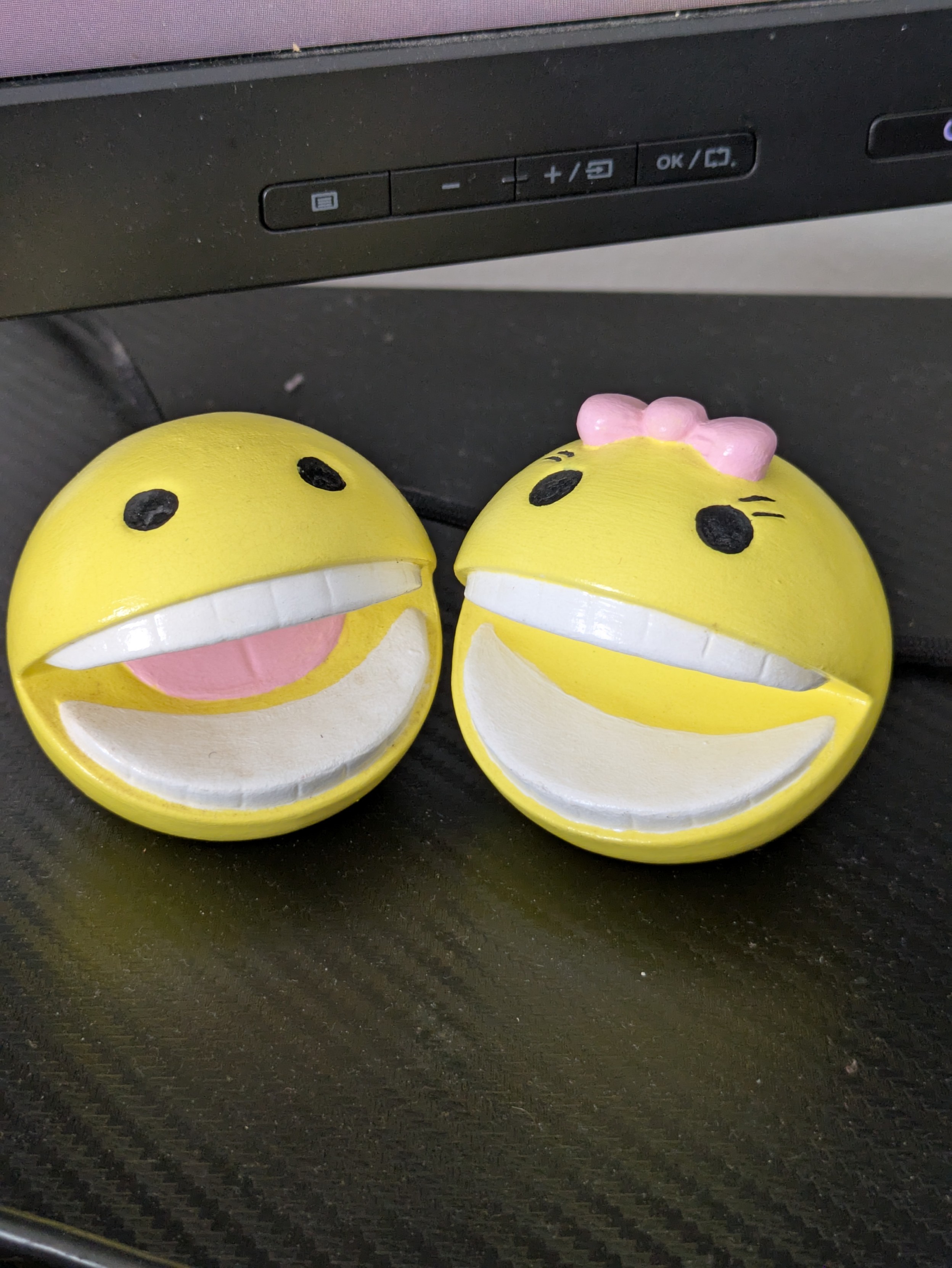 Pac-Man and Ms. Pac-Man figures sitting side by side on a desk with a monitor in the background. Each has creepy human-like teeth. Pac-Man has a tongue and Ms. Pac does not.