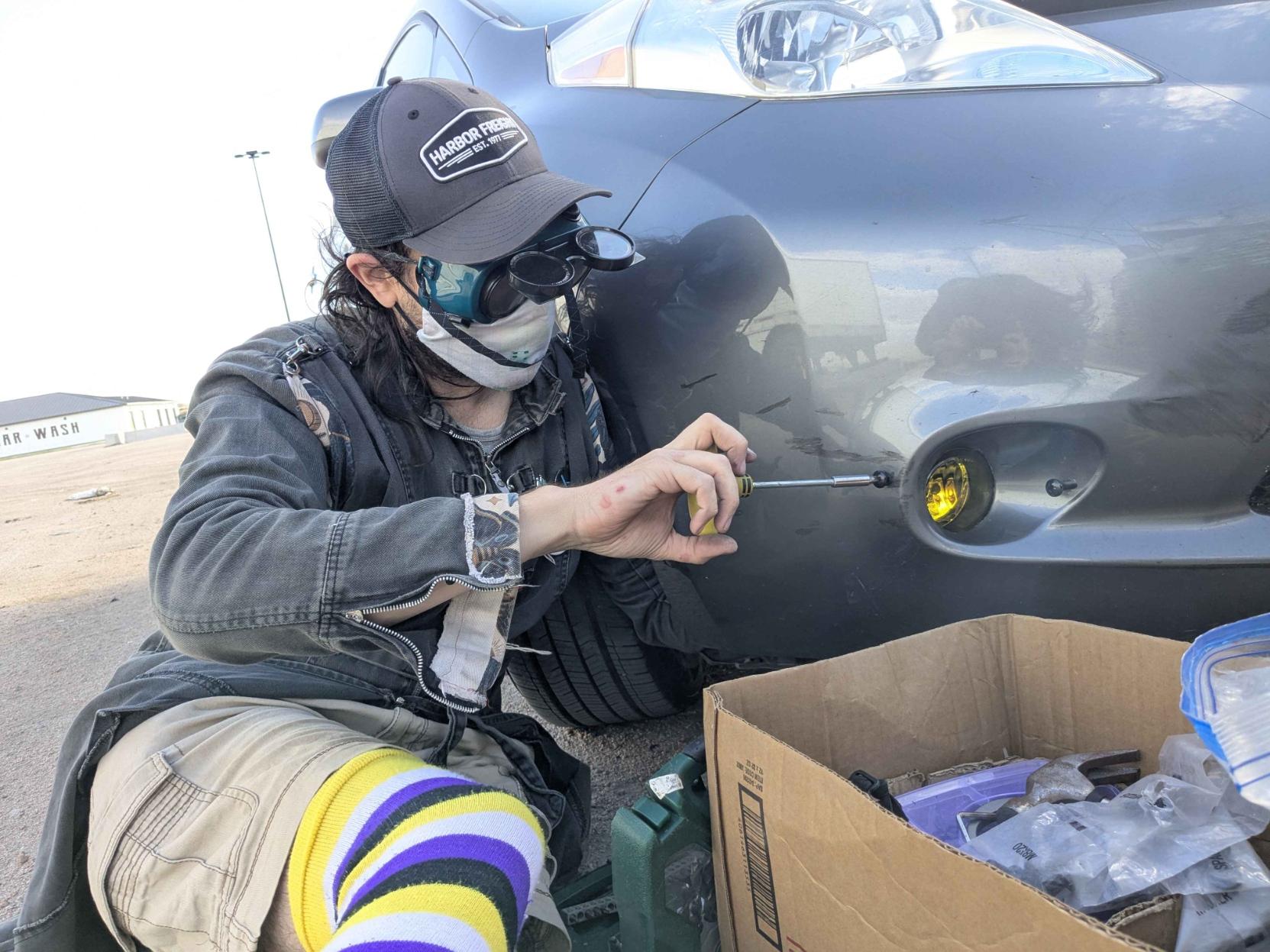 A non-binary person (Atha) sitting with hir left arm extended into the front corner of a grey Nissan Leaf bumper and hir right hand holding a screwdriver, tightening a screw to hold the fog light in place. The front wheel of the Nissan is steered outwards to facilitate access. A box of tools and parts is in the foreground at right of image. A green case of drill and screwdriver bits is just behind it in centre of image. Atha is sat on the sandy and dusty parking lot with buildings visible in the distance at far left, Atha is dressed in a Harbor Freight peak hat, hinged green welding goggles, a dust mask, a long black "Criminal Damage" denim coat modified with Etsy gothic print denim at the cuffs and shoulders, a grey t-shirt, beige 3/4 length shorts and a pair of non-binary flag pattern thigh high socks from Sock Dreams.